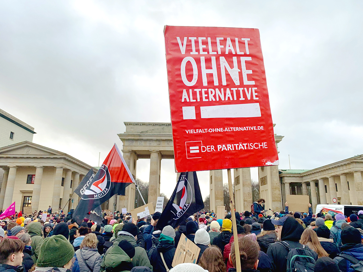 Vor dem Brandenburger Tor ist eine Menschenmenge zu sehen, die demonstriert. Im Vordergrund ein rotes Schild, auf dem in weißer Schrift steht: Vielfalt ohne Alternative! Der Paritätische